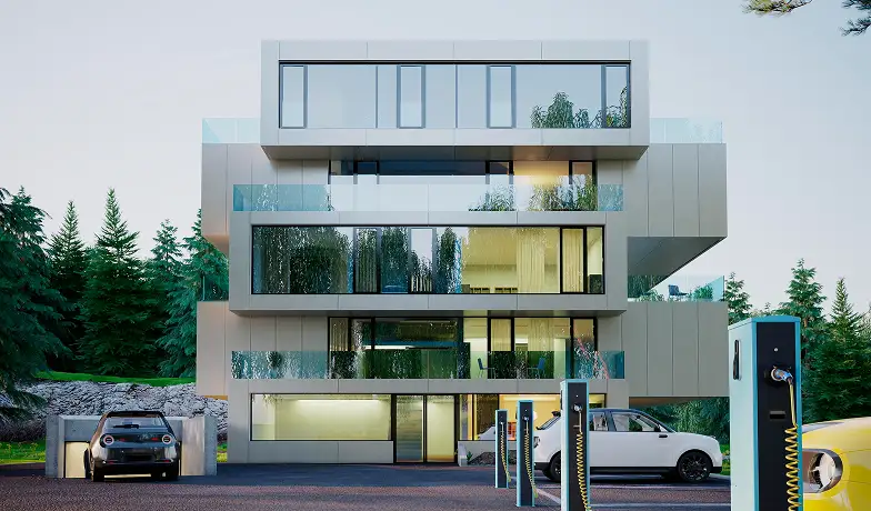 Modern, háromszintes épület nagy üvegablakokkal és geometrikus formatervezéssel, fákkal körülvéve. Előtte elektromos autók állnak, töltőállomásokkal.