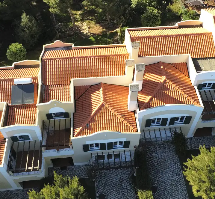 A dombtetőn álló, vörös cseréptetős házakat sűrű, zöld erdő és sziklás táj veszi körül. A látvány nyugodt, lágy fény és tiszta égbolt jellemzi.