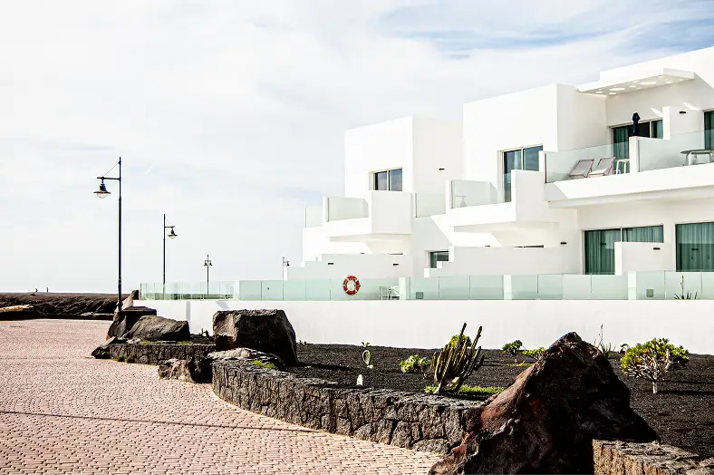 A modern, fehér épület erkélyeivel és üvegkorlátjaival a part menti sétányra néz. A sziklás kerttervezés és az utcai lámpák nyugodt, minimalista hangulatot teremtenek.