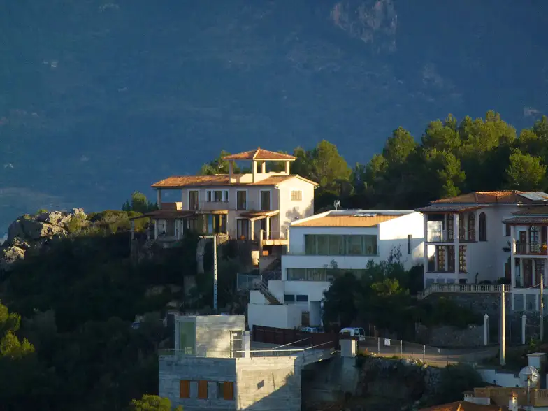 A dombtetőn álló, vörös cseréptetős házakat sűrű, zöld erdő és sziklás táj veszi körül. A látvány nyugodt, lágy fény és tiszta égbolt jellemzi.