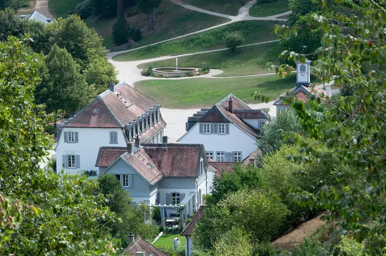 Fehér, történelmi villák csoportja vörös tetőkkel, buja zöld fák között megbújva, háttérben gondozott kerttel és ösvényekkel, amelyek nyugalmat árasztanak.