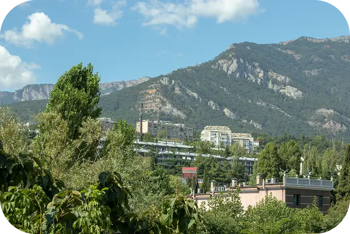 Gyönyörű kilátás egy buja, zöld tájra, magas hegyekkel a háttérben. A fák között megbújó épületek nyugodt, békés hangulatot árasztanak.