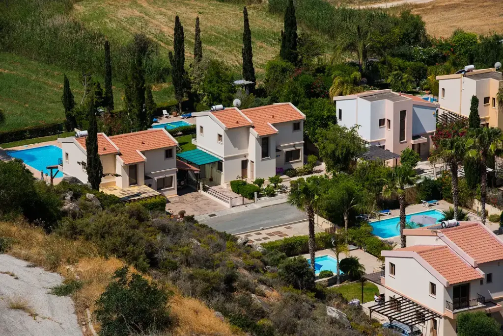 Légi felvétel egy békés lakónegyedről, modern házakkal, terrakotta tetőkkel, buja zöld kertekkel és kristálytiszta kék medencékkel, mezőkkel körülvéve.